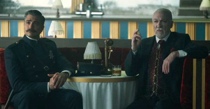 Jaime Camil în rolul sergentului Rivera și Patrick Page în rolul lui Octavius Kratt, stând la clubul Kratt, arătând viclean.'Schmigadoon'.