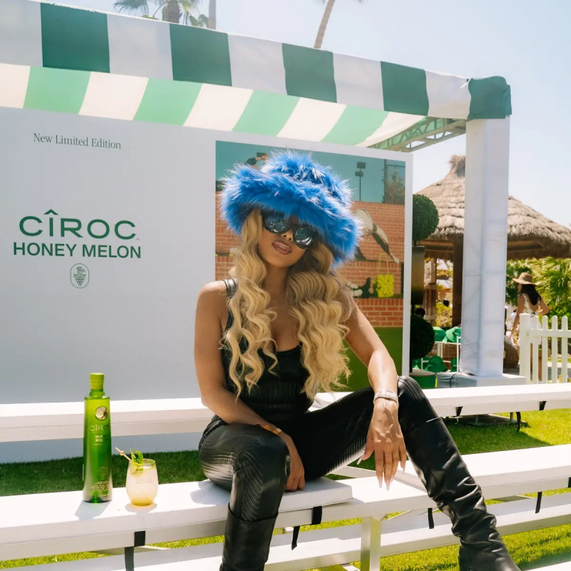 Lady London은 Circo 옆에 앉아 파란색 솜털 모자와 선글라스를 쓰고 있습니다.'s Honeydew Melon vodka.
