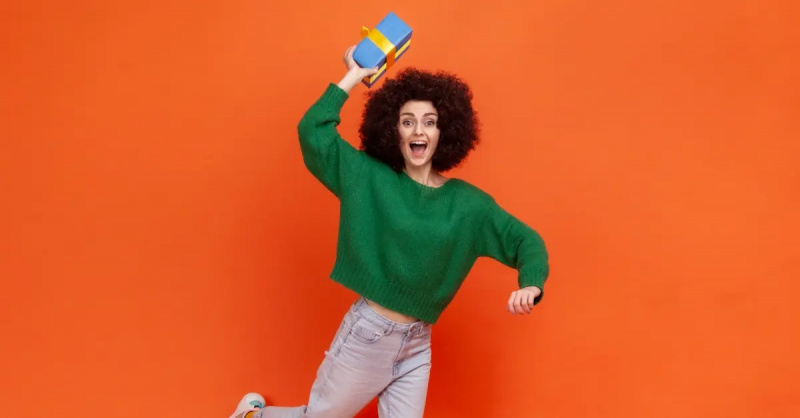 Babae na may berdeng sweater sa isang orange na background na may hawak na asul na kahon ng regalo.