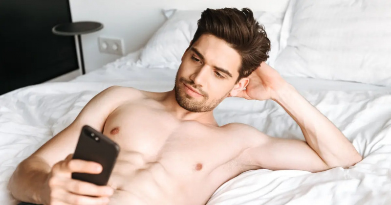 男性がベッドでシャツなしで携帯電話を見る。
