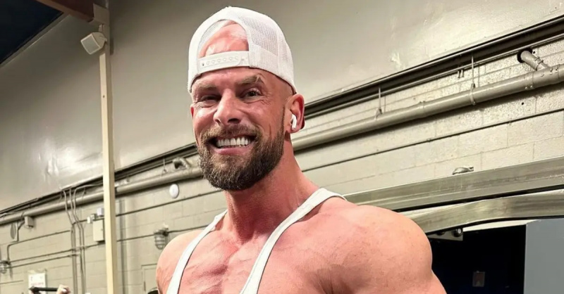 乔伊·斯沃尔(Joey swole)向后戴白色卡车司机帽和白色背心,在健身房微笑