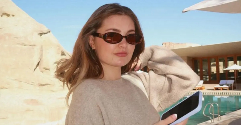 Aspyn Ovard na naka sunglass at sweater habang hawak ang telepono sa labas sa tabi ng pool