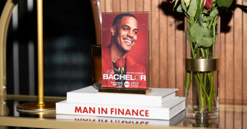 การ์ดภาพถ่ายของ'The Bachelor' star Grant Ellis sits on top of white books. The spine of the bottom book says "Man in Finance" in red coloring.
