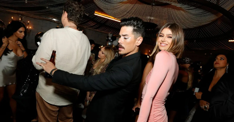 Tom Sandoval se uită în jos la o petrecere în timp ce Rachel Leviss pozează peste umărul ei, lângă spatele lui