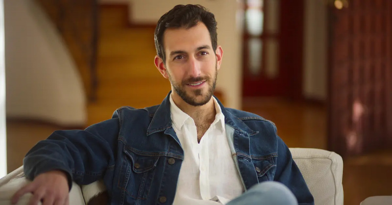 אורי מ'Jewish Matchmaking' sitting on a sofa, wearing collared shirt and denim jacket