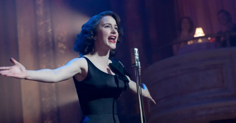 仪'Midge' Maisel (Rachel Brosnahan) in a black dress, delivers a comedy routine in front of a silver microphone