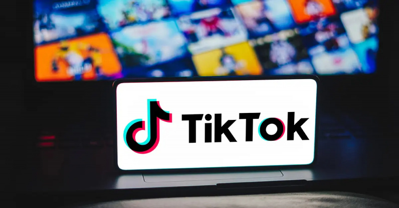 Логотип TikTok на экране телефона, за которым находится ноутбук.